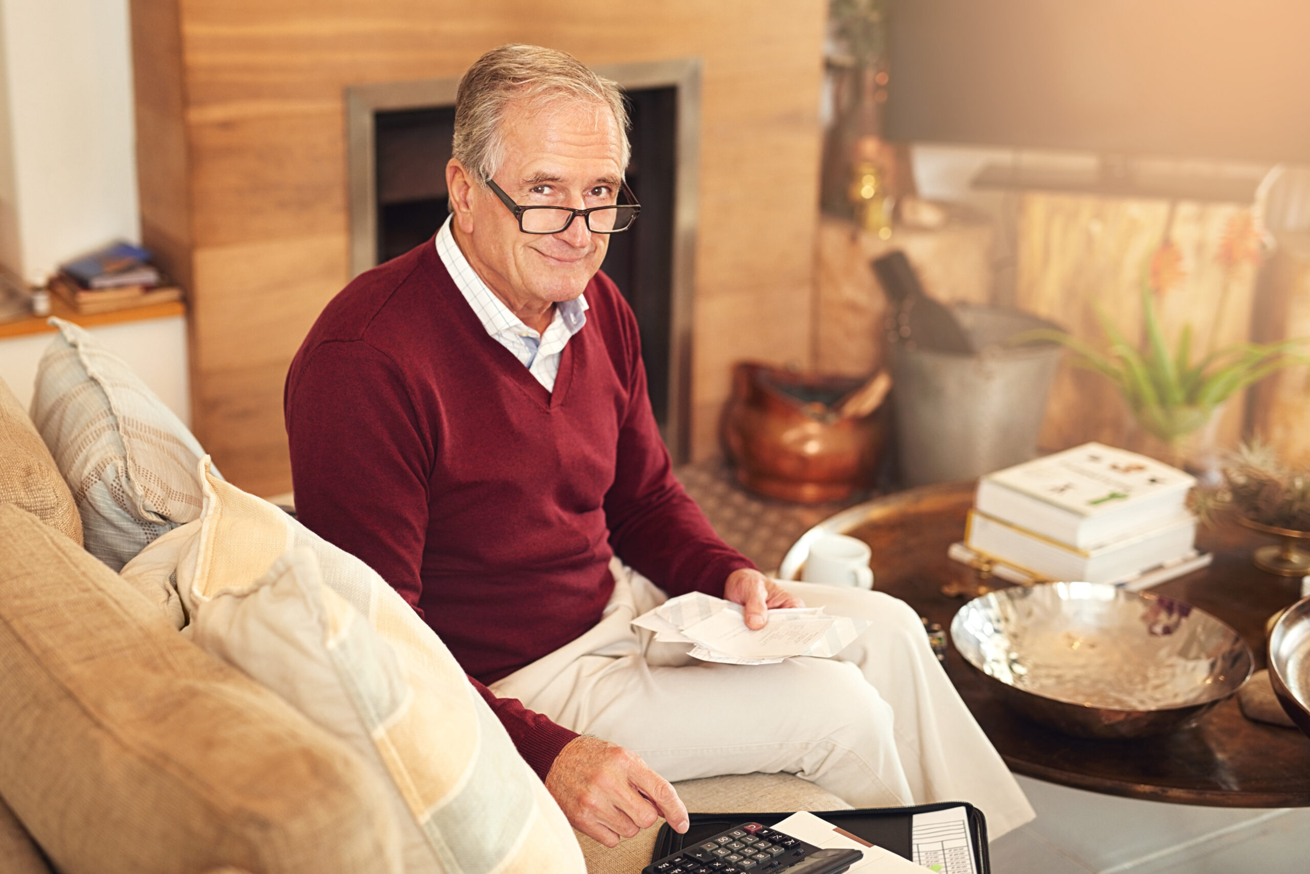 Senior man sitting on couch going through paperwork with calculator. 